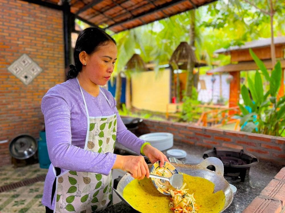 Cô Tư đổ bánh xèo vàng giòn trên chảo ở bếp ngoài trời tại Làng du lịch sinh thái Ông Đề