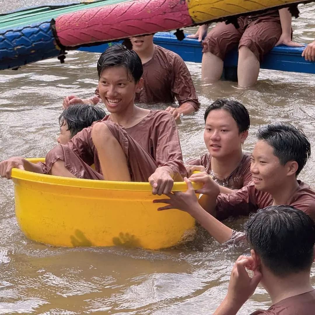 Bạn trẻ ngồi trong thau nhựa vàng giữa ao, các bạn đứng dưới nước bám thành thau cười đùa