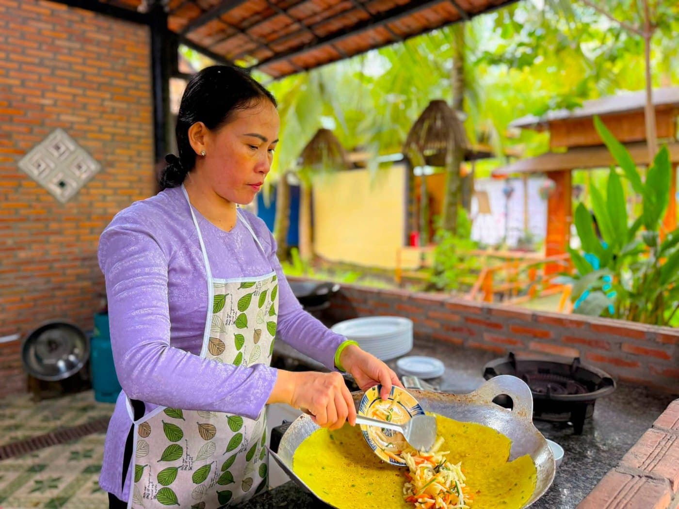 Cô Tư đổ bánh xèo vàng giòn trên chảo ở bếp ngoài trời tại Làng du lịch sinh thái Ông Đề