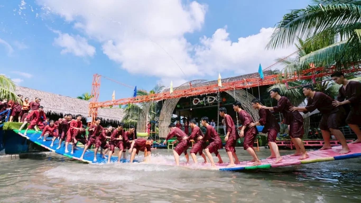 Đông đảo du khách áo nâu cùng nhún trên cây cầu nhiều màu bắc qua ao, người xem đứng dưới mái lá